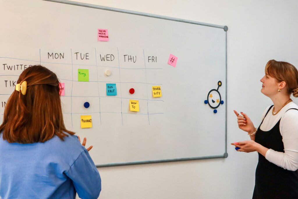 Two women checking a schedule made with sticky notes showing a week of work