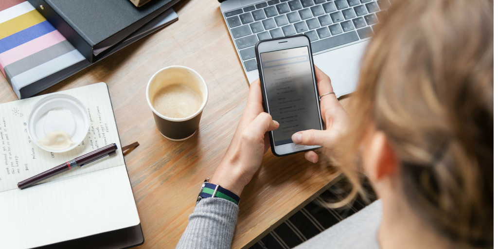 woman searching on her phone, at her desk with her laptop and some coffee